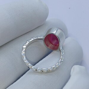 May include: A silver ring featuring a circular bezel setting with a vibrant pink stone. The band of the ring has a textured, notched edge design that runs along the entire circumference, providing a distinct metallic finish to the piece.