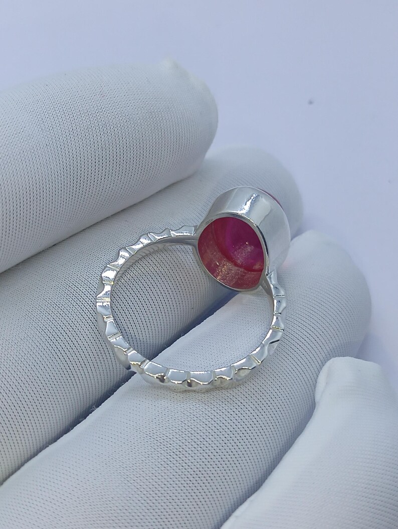May include: A silver ring featuring a circular bezel setting with a vibrant pink stone. The band of the ring has a textured, notched edge design that runs along the entire circumference, providing a distinct metallic finish to the piece.