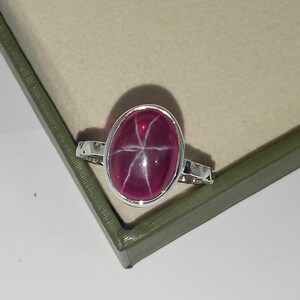 May include: A silver ring featuring a large, oval-shaped, deep pink star gemstone set in a smooth metal bezel. The gemstone displays a distinct six-rayed star pattern across its polished, cabochon-cut surface. The ring band has a textured, notched edge.