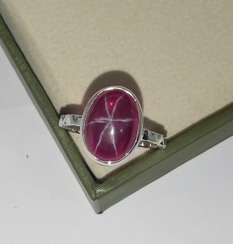 May include: A silver ring featuring a large, oval-shaped, deep pink star gemstone set in a smooth metal bezel. The gemstone displays a distinct six-rayed star pattern across its polished, cabochon-cut surface. The ring band has a textured, notched edge.