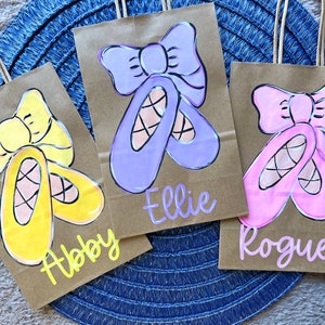 May include: Three brown paper gift bags with hand-painted ballet shoes and bows in yellow, purple, and pink. Each bag has a name written on it: Abby, Ellie, and Rogue. The bags have brown paper handles.