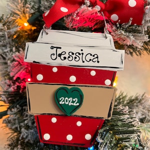 May include: A red and white polka-dot coffee cup ornament with a white lid featuring the name Jessica in black script. A tan sleeve wraps the center, adorned with a green heart displaying the year 2022. A red and white polka-dot ribbon bow hangs above.