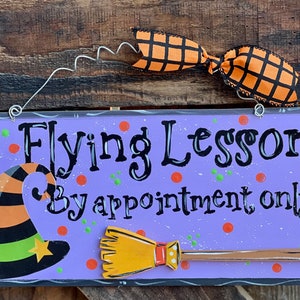 May include: A rectangular purple wooden sign with the black text "Flying Lessons By appointment only". The sign features a painted witch hat, a broomstick, and orange and green polka dots. An orange and black plaid ribbon is attached to the top.