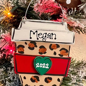 May include: A flat, coffee cup-shaped ornament featuring a tan background with a black and brown leopard print pattern. The cup has a white lid with the name Megan written in black, a red sleeve, and a green heart with the year 2022 in white.