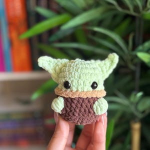 May include: A crocheted figure of a character with large ears, green skin, and black eyes. The figure is wearing a brown robe and has a tan collar. The figure is being held in a person's hand. The background is blurred with green foliage.