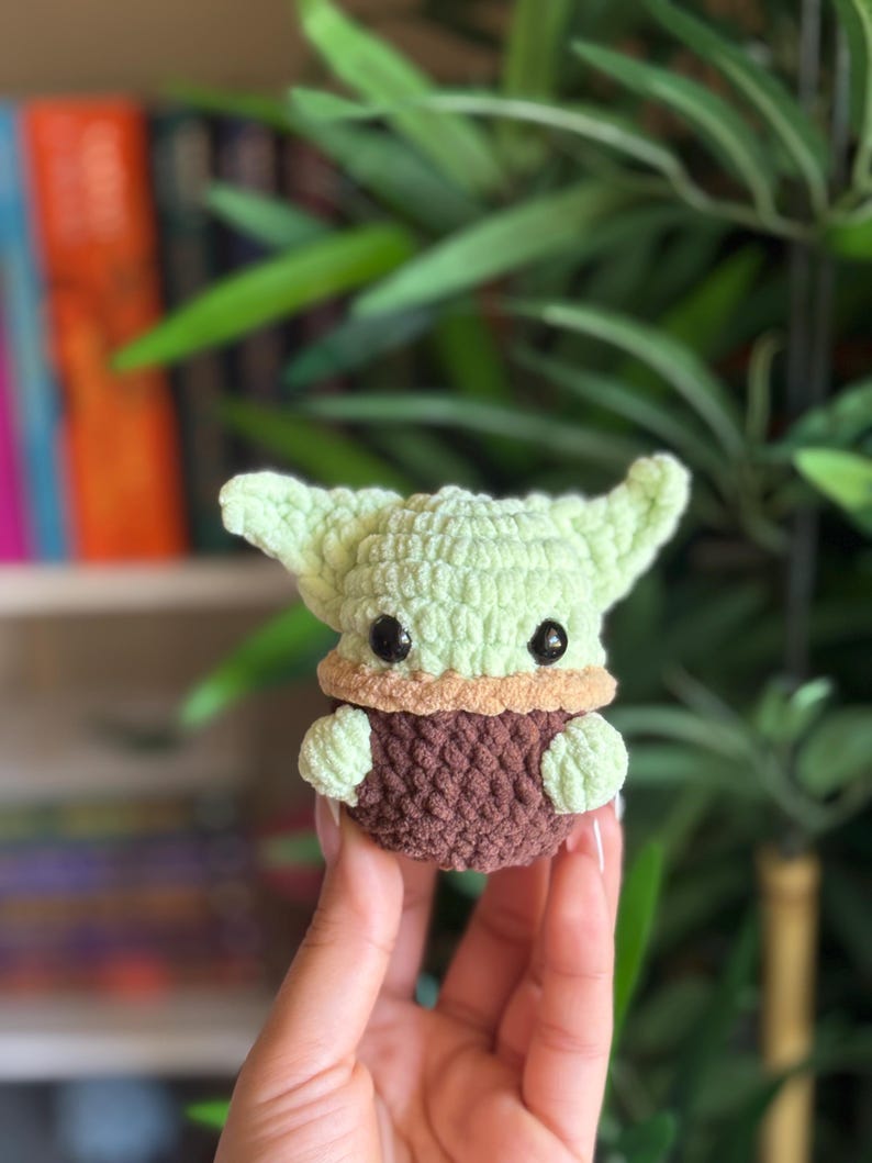May include: A crocheted figure of a character with large ears, green skin, and black eyes. The figure is wearing a brown robe and has a tan collar. The figure is being held in a person's hand. The background is blurred with green foliage.