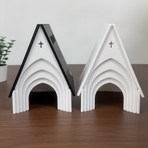 Op de afbeelding: Twee A-vormige structuren in de vorm van kerkgebouwen, één met een zwart dak en witte muren, de andere volledig wit. Beide hebben gewelfde ingangen met getrapte lijsten en een kleine kruisvormige uitsparing bij de nok.