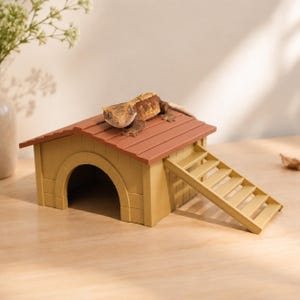以下が含まれることがあります： 茶色の屋根とアーチ型の入り口を持つ、小さな家のような形のベージュ色の爬虫類用シェルター。茶色とベージュのまだら模様の皮膚を持つクレステッドゲッコーが屋根の上に休んでいる。構造物の側面から屋根へと続く階段が付いている。