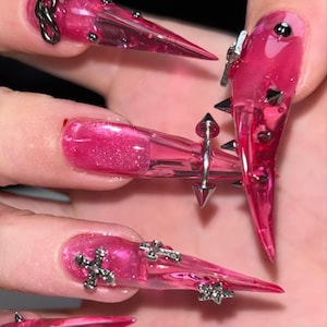 Può includere: Primo piano di unghie acriliche lunghe, appuntite e rosa. Le unghie sono decorate con borchie argentate, croci e abbellimenti a forma di stella. Alcune unghie presentano un design ad anello argentato. Le unghie hanno una finitura lucida.