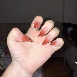 Könnte beinhalten: Eine Hand mit langen, spitzen Fingernägeln im Ombre-Stil. Die Nägel gehen von einer klaren Basis zu einem tiefen Rot an den Spitzen über. Ein silberner Ring befindet sich am Ringfinger. Die Nägel haben ein scharfes, krallenartiges Aussehen.