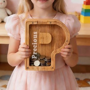 May include: A wooden piggy bank in the shape of the letter "P" with the word "Precious" printed on it. The bank is filled with coins. The girl is wearing a pink dress.