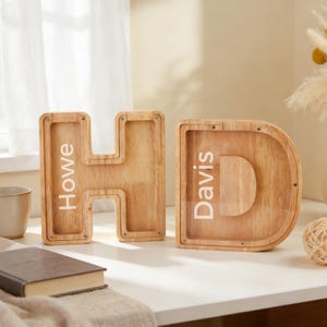 Puede incluir: Dos huchas de madera con forma de letra. La letra "H" tiene el nombre "Howe" impreso, y la letra "D" tiene el nombre "Davis" impreso. Los bancos están hechos de madera de color claro y tienen un panel frontal transparente.