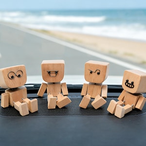 May include: Four small wooden robot figurines with articulated limbs and block-shaped heads. Each figure features a unique facial expression created with thin black wire, including circular eyes, a rectangular mouth, a curved line, and a wide grin.