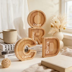 Puede incluir: Huchas de madera con forma de letras que deletrean "CORD" con nombres inscritos. Las letras son de color marrón claro con un frente transparente. Una taza beige, libros y objetos decorativos están en el fondo. La letra "C" tiene inscrita "Caine".
