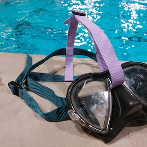 Könnte beinhalten: Eine schwarze Tauchmaske mit klaren Gläsern und einem schwarzen Rahmen. Die Maske verfügt über ein verstellbares, blaugrünes Riemensystem, das um den Kopf verläuft, ergänzt durch einen hellvioletten Stoffriemen an der Oberseite des Rahmens.