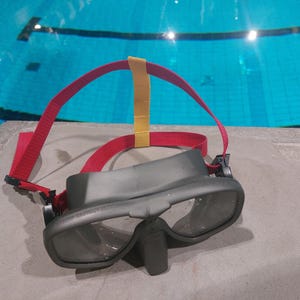 Könnte beinhalten: Graue Taucherbrille mit klaren Gläsern und einem schwarzen Nasenstück. Die Brille hat ein rotes Band mit einem gelben Band oben. Die Brille liegt auf einer Betonoberfläche mit einem Schwimmbecken im Hintergrund.