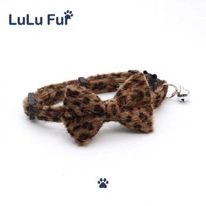 Puede incluir: Un collar para mascotas con estampado de leopardo marrón y negro con pajarita a juego. El collar tiene una pequeña campana plateada adjunta y hebillas de plástico negro. La marca "LuLu Fur" está impresa en azul.