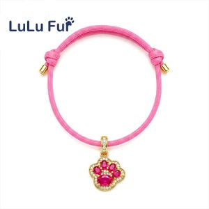 May include: Pink adjustable pet bracelet with a gold-coloured paw print charm. The charm is encrusted with red and clear gemstones. The bracelet has gold-coloured metal accents and the brand name "LuLu Fur" in the upper left corner.