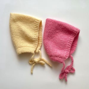Pode incluir: Dois gorros de malha, um num amarelo pálido suave e o outro num rosa vibrante. Cada gorro apresenta um padrão de malha texturizado, uma borda canelada em torno da abertura facial e laços de fio a condizer atados na parte inferior.