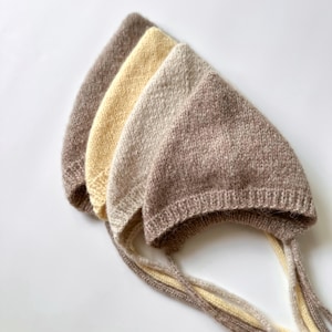 Può includere: Un set di quattro cuffiette lavorate a maglia nei toni del marrone, crema e giallo pallido. Ogni cuffietta presenta una parte superiore a punta, un bordo a coste attorno all'apertura per il viso e due lunghi lacci lavorati a maglia agli angoli.