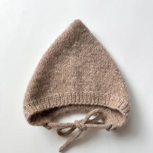May include: A light brown, pointed knit bonnet featuring a ribbed trim along the bottom edge and two matching knitted ties that are knotted together at the front. The hat is crafted from a soft, textured yarn with a subtle heathered appearance.