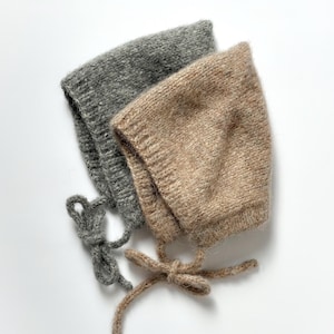 Op de afbeelding: Twee gebreide baby mutsjes. Eén is lichtgrijs van kleur en de andere is lichtbruin van kleur. Beide mutsjes hebben strikjes bij de kin. De mutsjes zijn gemaakt van een zacht, getextureerd garen.