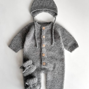 Peut inclure: Ensemble de vêtements pour bébé en tricot gris, comprenant une combinaison boutonnée, un bonnet assorti avec des liens et une paire de chaussons. L'ensemble est fait d'un fil doux et texturé et comporte des boutons en bois. L'ensemble est conçu pour les nouveau-nés.