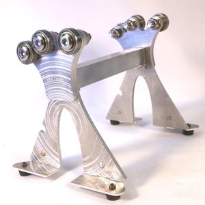 以下が含まれることがあります： 複数のローラーベアリングを備えた、銀色の金属製工具スタンド。スタンドは、湾曲した切り欠きと黒いゴム製の足を備えた、頑丈で角張ったデザインです。「HPG Tools」の文字が側面に表示されています。