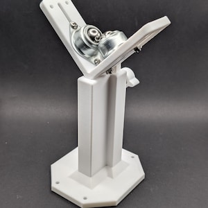 May include: A white, 3D-printed device with a hexagonal base and a vertical support structure. Two metal ball bearings are mounted on angled white arms, secured with screws. The device is designed for mechanical use.