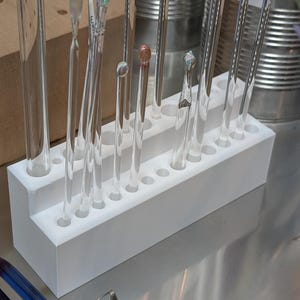 Puede incluir: Un soporte blanco de dos niveles con múltiples agujeros, lleno de varias herramientas de vidrio transparente. Algunas herramientas tienen extremos redondeados o puntiagudos. El fondo incluye recipientes metálicos y un bloque de madera, lo que sugiere un taller o laboratorio.