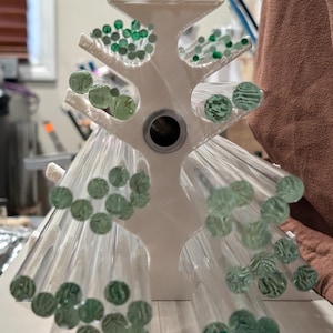 Puede incluir: Un expositor blanco en forma de árbol que contiene múltiples varillas de vidrio transparente con acentos circulares verdes. Las varillas están dispuestas para parecerse a ramas, con los acentos verdes que imitan las hojas. El soporte tiene un agujero central.