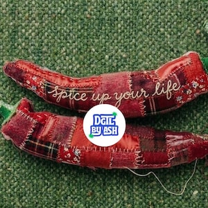 May include: Two handmade red chili pepper-shaped fabric ornaments with green stems. The peppers are made of patchwork fabric with the words "spice up your life" embroidered on one. The background is green.