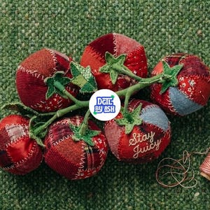 Puede incluir: Un grupo de tomates de tela hechos a mano con tallos y hojas verdes, sobre una superficie verde texturizada. Los tomates son rojos con detalles de patchwork, y uno tiene el texto "Stay Juicy". También hay una bobina de hilo.