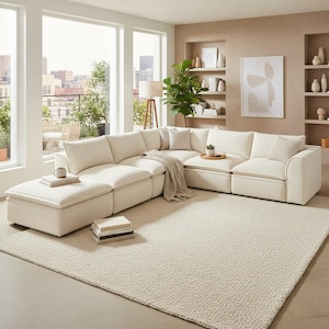 May include: A cream-colored sectional sofa with a corduroy texture, arranged in an L-shape, dominates a bright living room. The sofa is paired with a matching ottoman and a textured rug. Pillows and a throw blanket add comfort. A coffee table holds a tray with a plant.