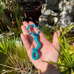 Op de afbeelding: Een turquoise hangerketting met een serpentijnontwerp. De hanger wordt in een hand gehouden en de ketting heeft een kralenstreng met blauwe en heldere kralen. De achtergrond omvat groen en een waterpartij.
