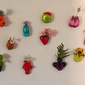May include: A collection of ten small, colorful ceramic wall-mounted vases in various shapes, including heart, pear, and abstract forms. Each vase features a glossy finish in colors like red, green, orange, blue, and purple, holding small floral stems.
