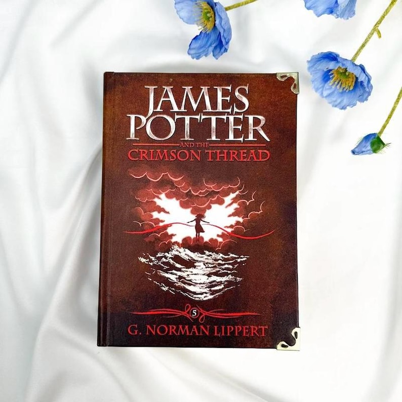 May include: A hardcover book titled "JAMES POTTER and the CRIMSON THREAD" with a dark red cover. The cover features a silhouette of a person and a red cloud design. The author's name, G. Norman Lippert, is at the bottom. Decorative gold corners and blue flowers are in the background.