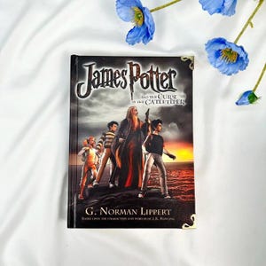 May include: A hardcover book titled "James Potter and the Curse of the Gatekeeper" by G. Norman Lippert. The cover features illustrations of characters and a wizard against a sunset backdrop. The book is decorated with gold corner accents.