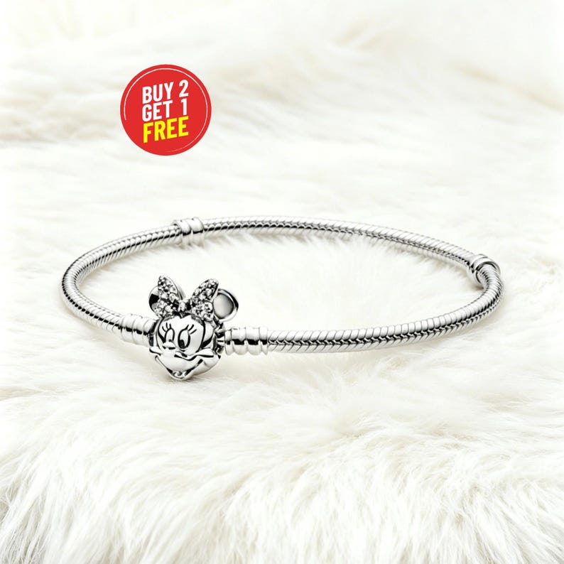 May include: A silver-colored bracelet with a Minnie Mouse charm. The charm features a bow and Minnie's face. The bracelet is on a white, fluffy surface. A red circle with the text "BUY 2 GET 1 FREE" is in the upper left corner.