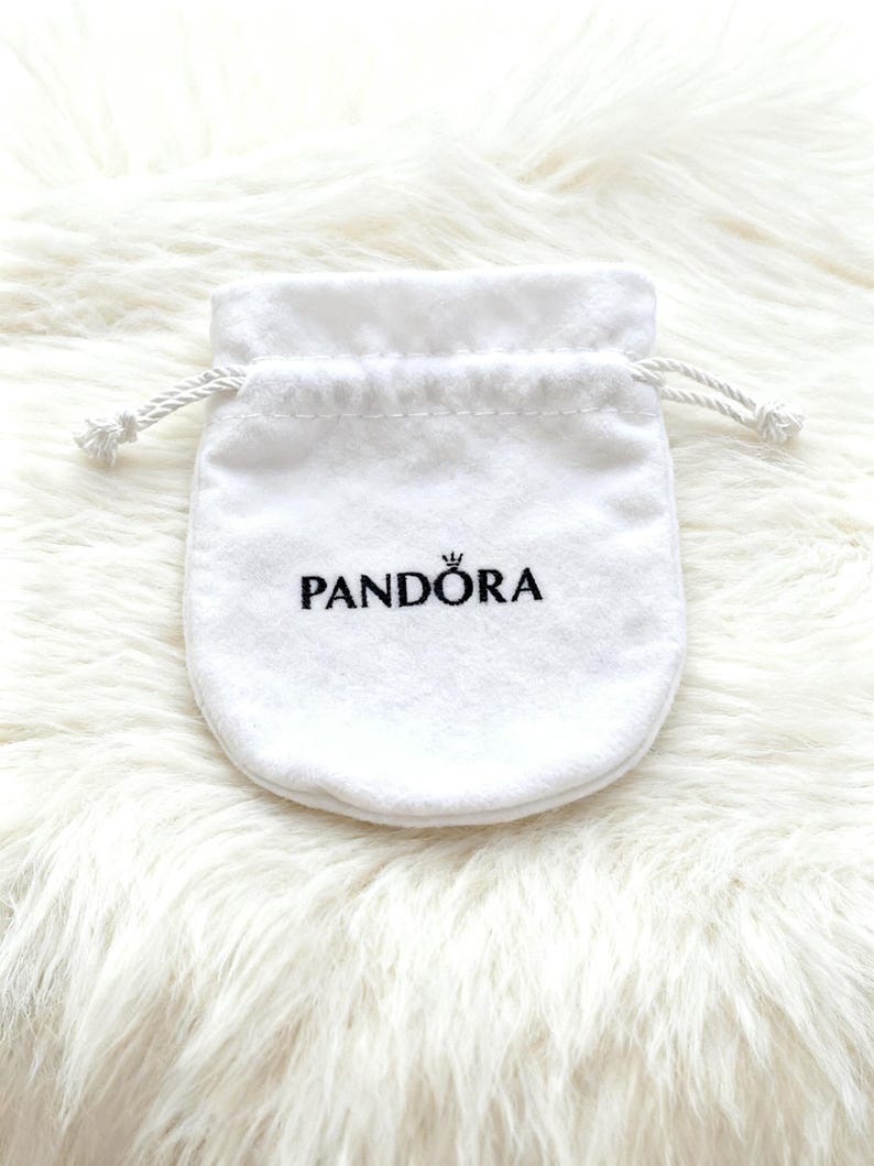 May include: A white, drawstring jewelry pouch with the brand name "PANDORA" printed in black. The pouch is made of a soft, velvety material and is set against a fluffy, white background.