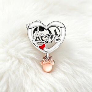 May include: A silver charm with a heart-shaped design. The word "Love" is written across the heart. A small red heart and a rose gold Minnie Mouse charm dangle below. The charm is set against a white, fluffy background.