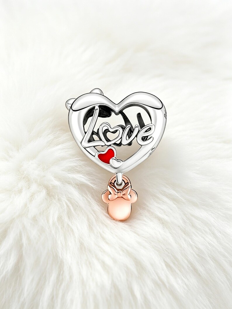 May include: A silver charm with a heart-shaped design. The word "Love" is written across the heart. A small red heart and a rose gold Minnie Mouse charm dangle below. The charm is set against a white, fluffy background.
