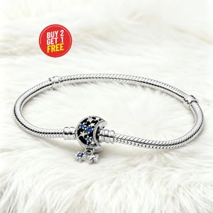May include: A silver bracelet with a charm featuring a crescent moon design with blue accents and dangling star charms. The bracelet is on a white, fluffy surface. A red circle with the text "BUY 2 GET 1 FREE" is in the upper left corner.