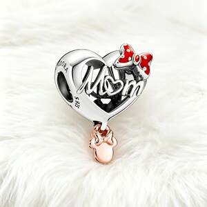 May include: A silver charm with the word "Mom" in script, shaped like a heart. A red and white polka dot bow is on the top right. A rose gold Minnie Mouse charm hangs below. The charm is on a white, fluffy surface.