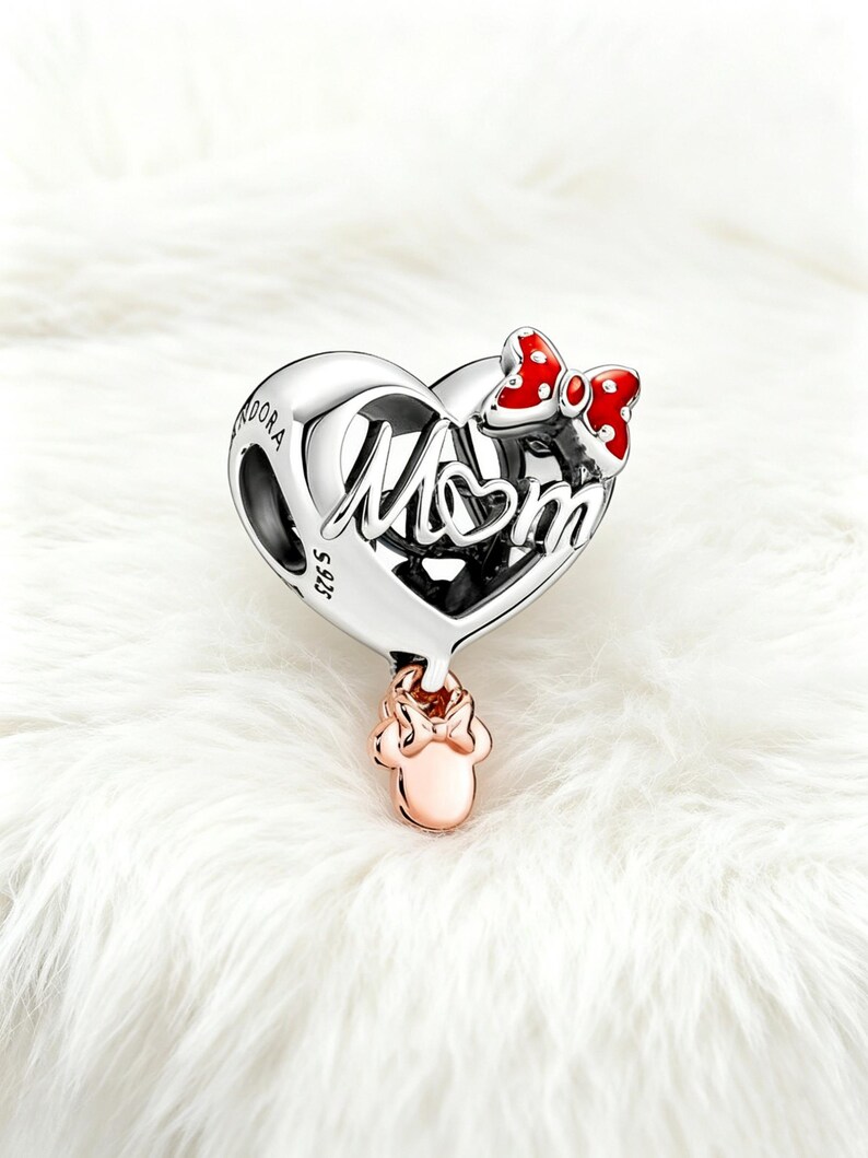 May include: A silver charm with the word "Mom" in script, shaped like a heart. A red and white polka dot bow is on the top right. A rose gold Minnie Mouse charm hangs below. The charm is on a white, fluffy surface.
