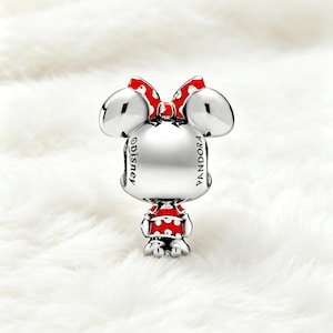 May include: A silver charm featuring a cartoon character with large ears and a red bow with white polka dots. The character wears a red and white polka dot outfit. The charm has the words "Disney" and "Pandora" engraved on it.