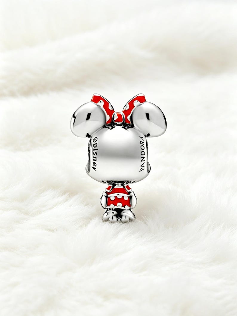 May include: A silver charm featuring a cartoon character with large ears and a red bow with white polka dots. The character wears a red and white polka dot outfit. The charm has the words "Disney" and "Pandora" engraved on it.