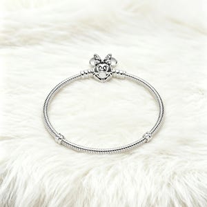 May include: A silver bracelet featuring a Minnie Mouse charm. The charm has a bow and a smiling face. The bracelet is made of a flexible, snake-like chain. The bracelet is displayed on a white, fluffy surface.