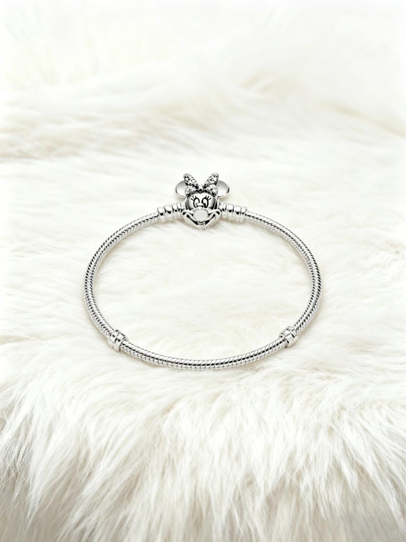 May include: A silver bracelet featuring a Minnie Mouse charm. The charm has a bow and a smiling face. The bracelet is made of a flexible, snake-like chain. The bracelet is displayed on a white, fluffy surface.