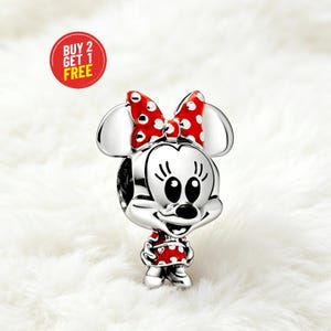 May include: A silver charm featuring Minnie Mouse with a red bow and dress, both with white polka dots. The charm has large ears and a smiling face. A red circle with the text "BUY 2 GET 1 FREE" is in the background.